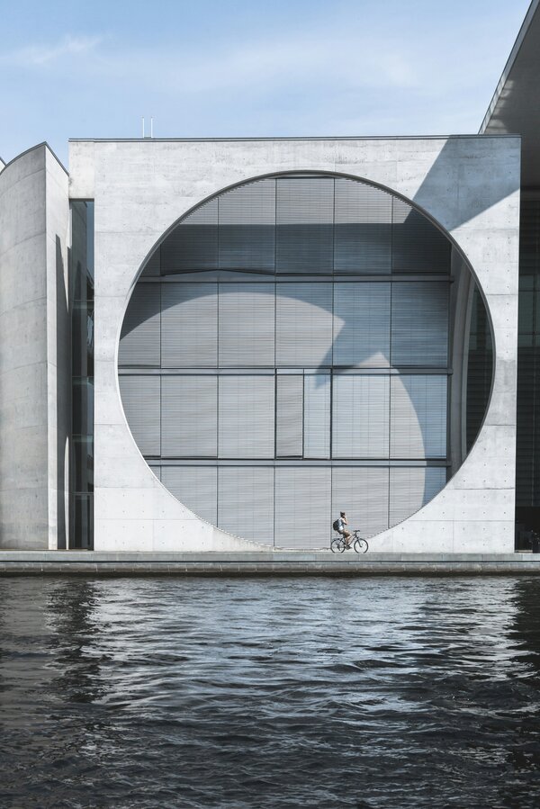 Photo urbaine en couleur, montrant un bâtiment carré dans lequel se trouve une ouverture ronde et des fenêtres rectangulaires, devant un point d'eau. Un cycliste roule au pied du bâtiment