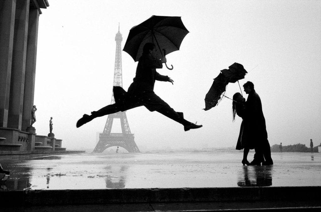 Photo noir et blanc très connue d'Elliott Erwitt représentant un homme sautant parapluie à la main, juste à côté d'un couple enlacé ayant chacun leur parapluie emporté par le vent (initiationphoto.com)