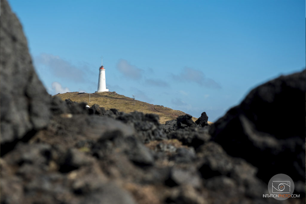 Photo d'une phare en haut d'une coline avec un cadre naturel créé par des rochers à l'avant-plan (initiationphoto)