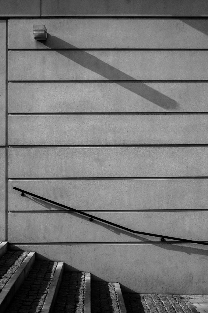 Photo en Noir et Blanc d'un mur avec des escaliers, une lampe, une rampe et des ombres portées