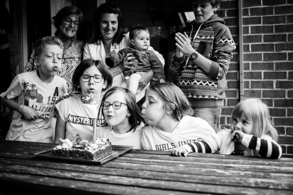 Photo en noir et blanc d'un petit groupe de personnes, majoritairement des enfants, dont quatre qui soufflent les bougies d'une gâteau